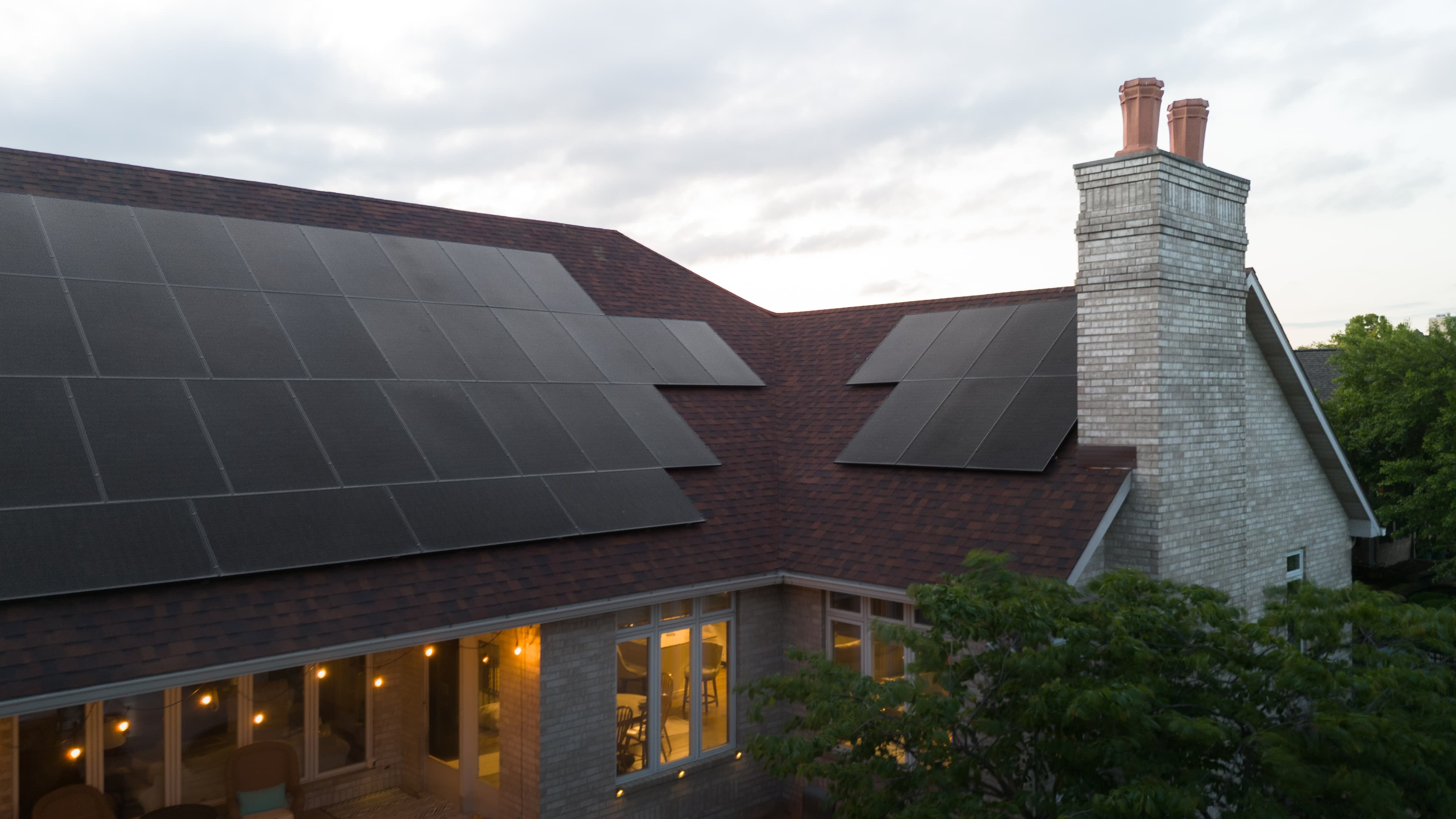 Residential rooftop with large solar panel array at dusk, warm interior lights visible through windows